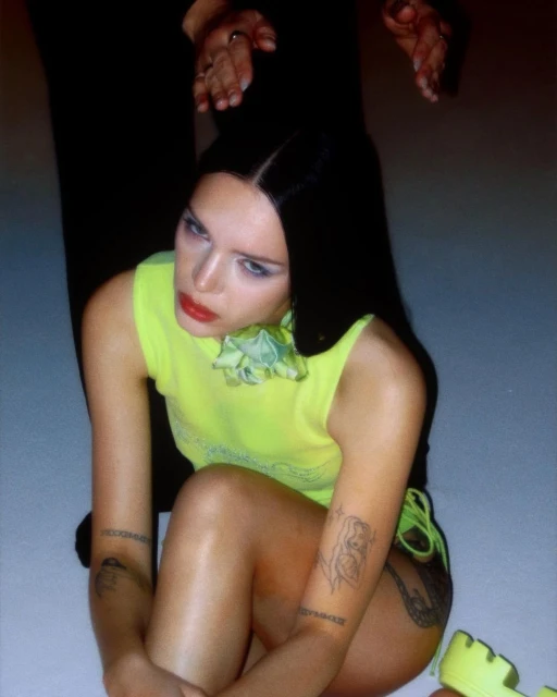 Young woman with dark hair and tattoos posing in a neon yellow top and matching platform shoes, sitting indoors with dramatic lighting.