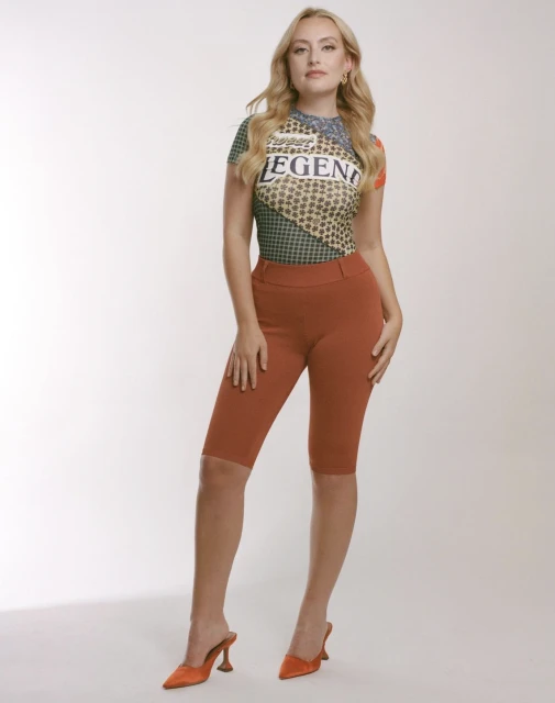 Full-length shot of a young woman posing in rust-colored shorts, a patterned top, and high heels against a white background in a photo studio.