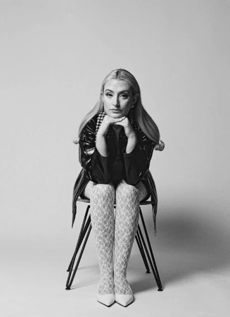 Blonde woman wearing patterned tights and a dark jacket seated on a chair against a plain seamless background.