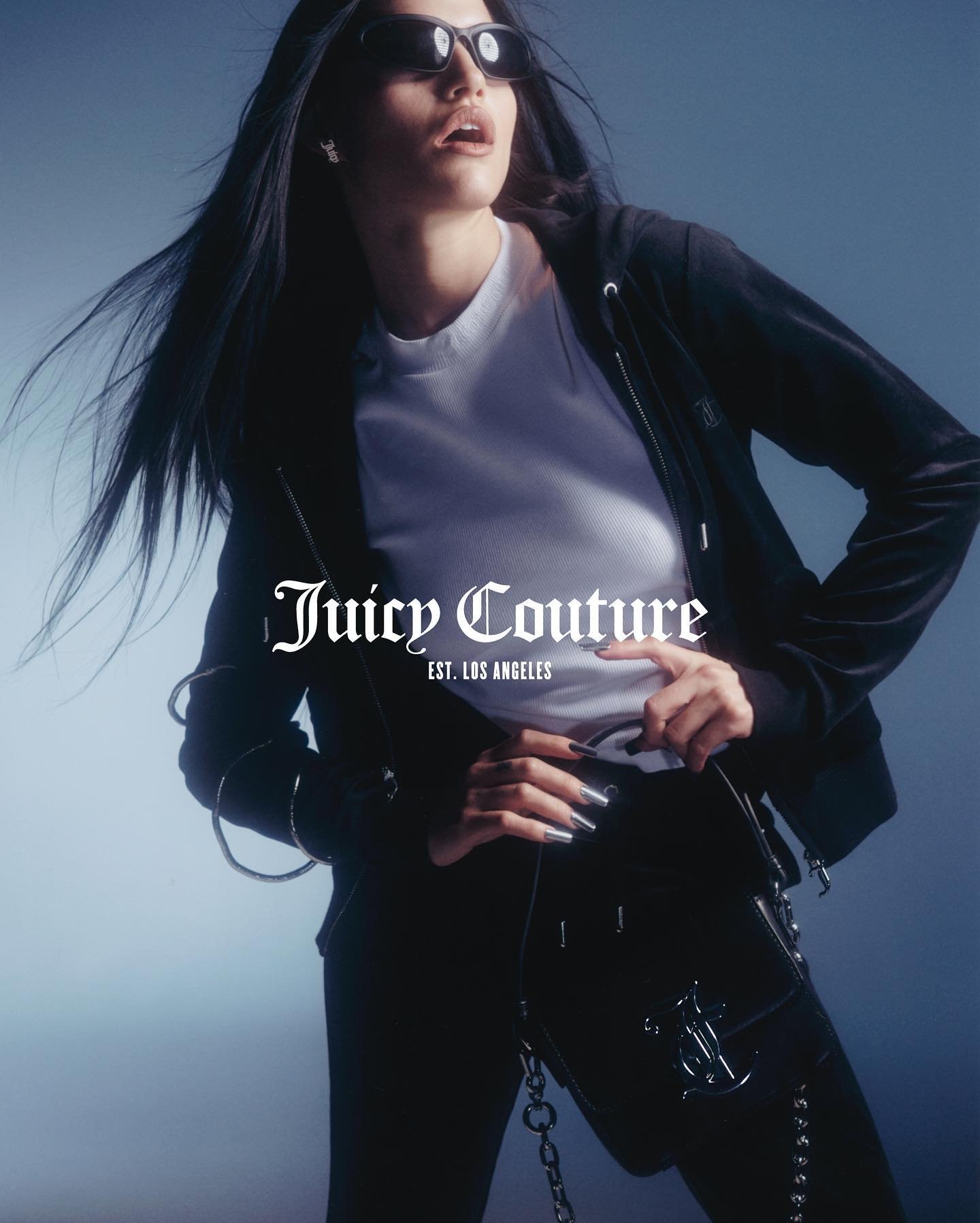 A woman poses in a black Juicy Couture tracksuit with sunglasses, showcasing fashion in a studio with soft lighting and a gradient backdrop.