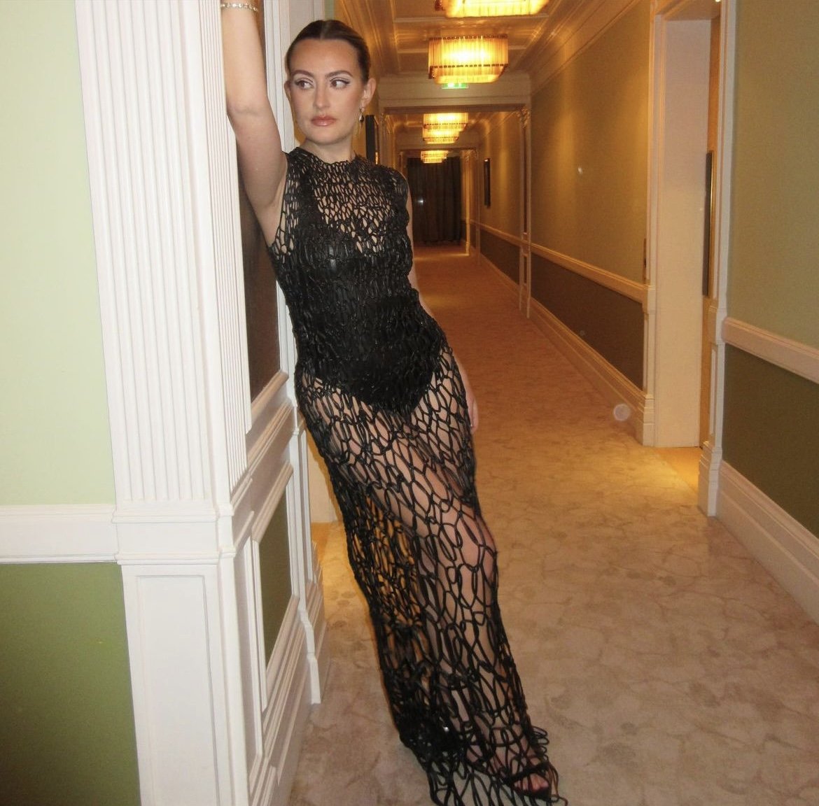 A woman in a black netted dress leans against a hallway wall, posing for a fashion shot with warm lighting in an indoor setting.