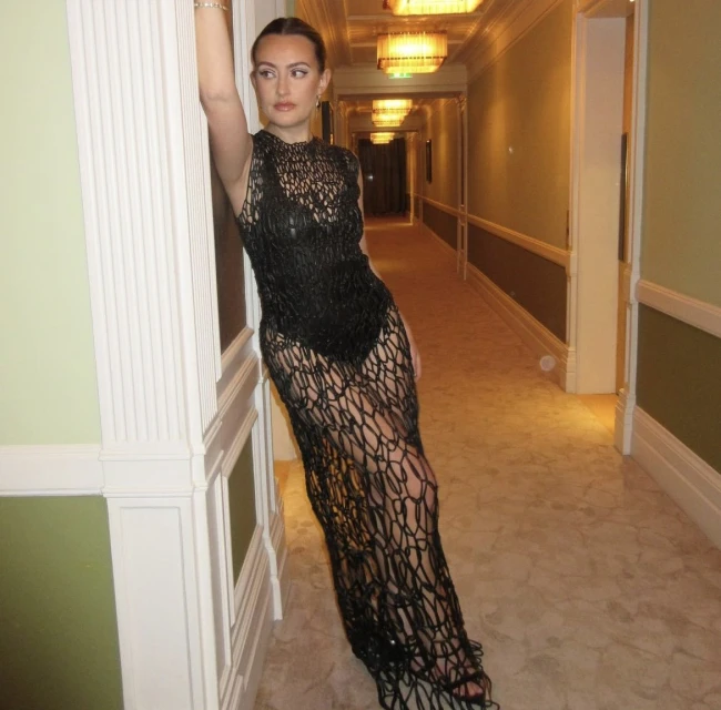 A woman in a black netted dress leans against a hallway wall, posing for a fashion shot with warm lighting in an indoor setting.