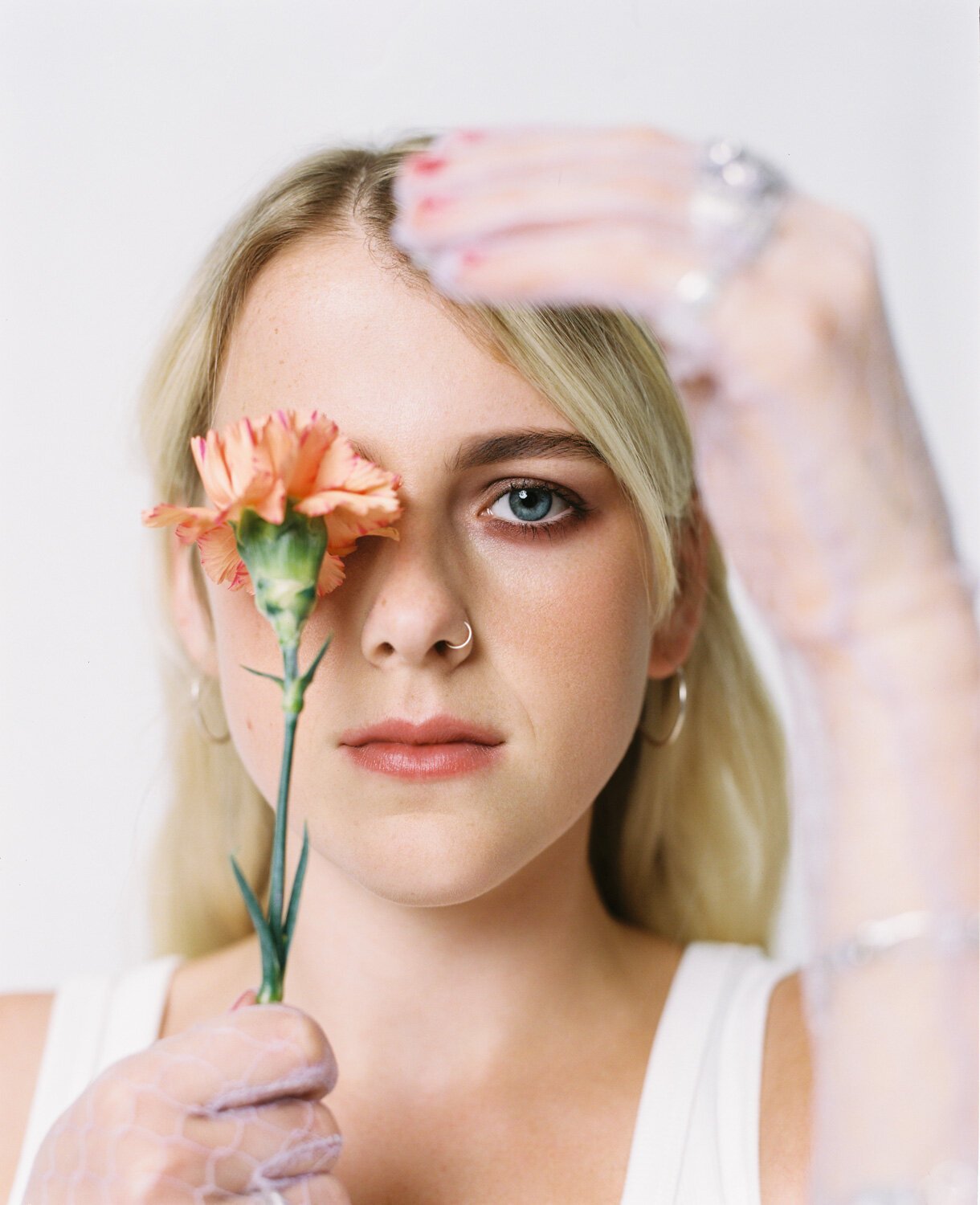 Close-up portrait of a blonde woman with a nose ring holding an orange carnation over one eye, wearing sheer gloves in a white setting.