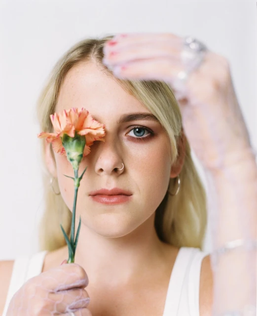 Close-up portrait of a blonde woman with a nose ring holding an orange carnation over one eye, wearing sheer gloves in a white setting.