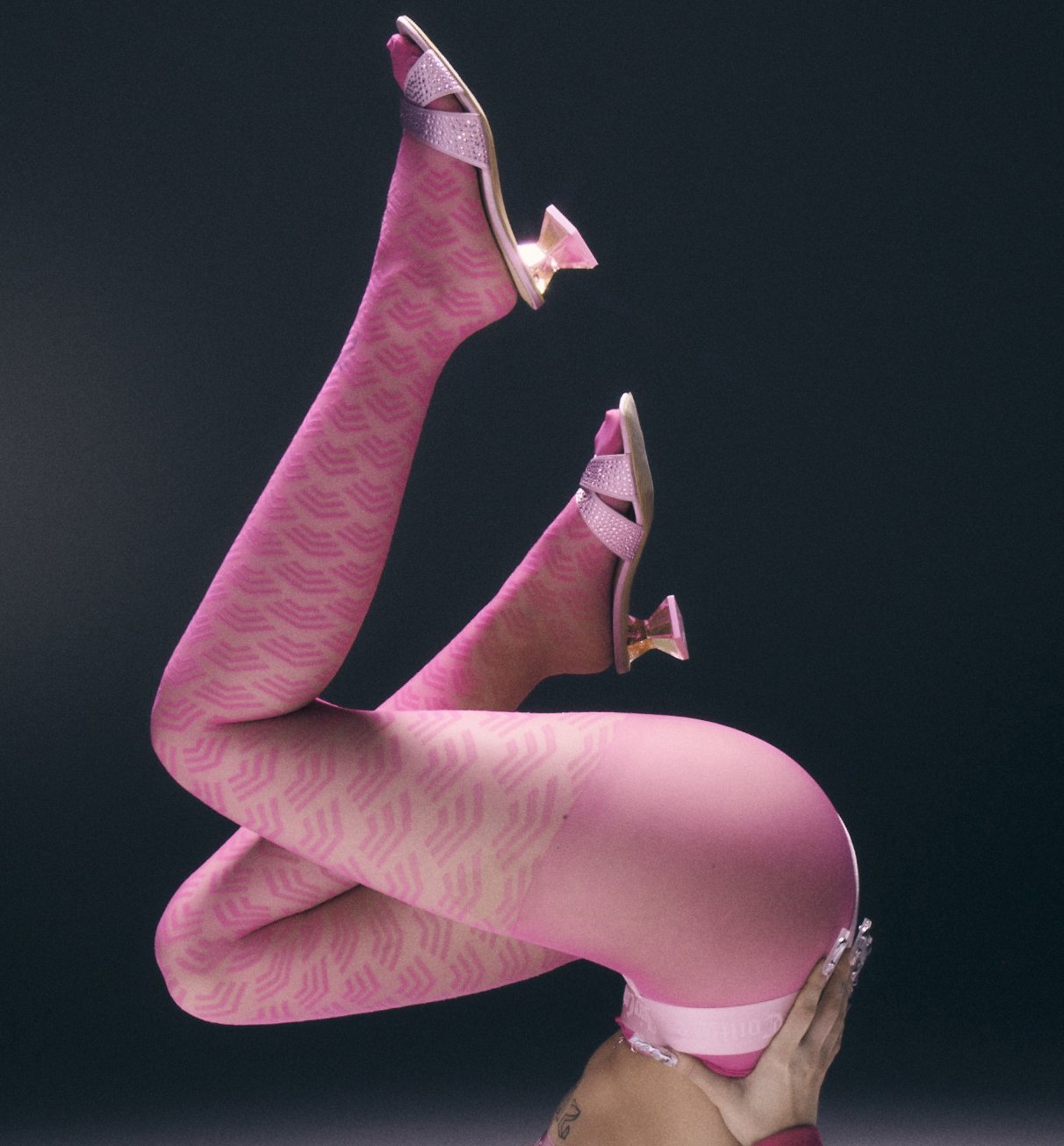 Elevated studio shot of a person with bent legs wearing pink patterned tights and high heels against a dark gray background.