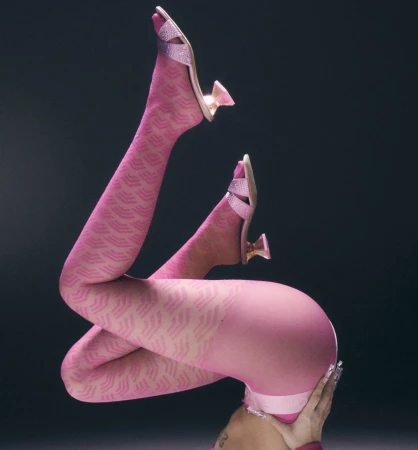 Elevated studio shot of a person with bent legs wearing pink patterned tights and high heels against a dark gray background.