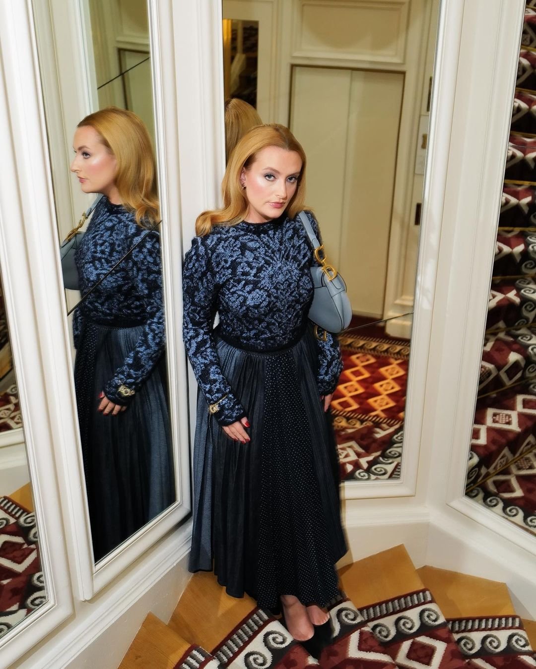 A woman in a blue patterned dress stands on a carpeted staircase, posing in front of a mirror in a hallway with neutral-toned walls.