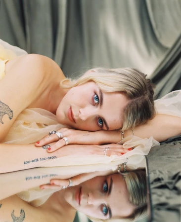 Blonde woman with blue eyes lying on light fabric resting chin on folded hands, reflected in a mirror below with visible tattoos.