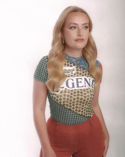 Blonde woman with patterned top and red pants in studio shot. Posing woman with soft facial expression, blonde wavy hair, indoor setting.