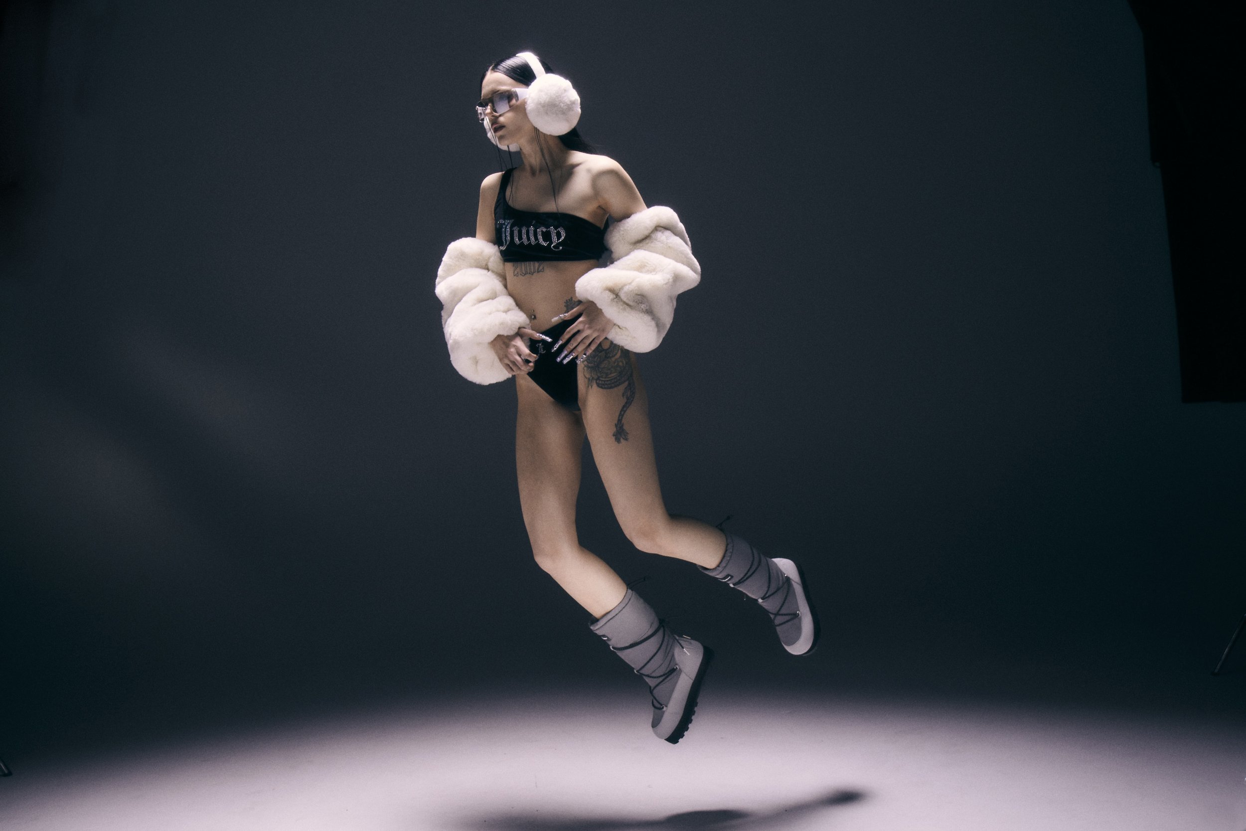 A woman poses in a photo studio wearing a bra top, briefs, earmuffs, and boots; full figure with a studio spotlight in the background.