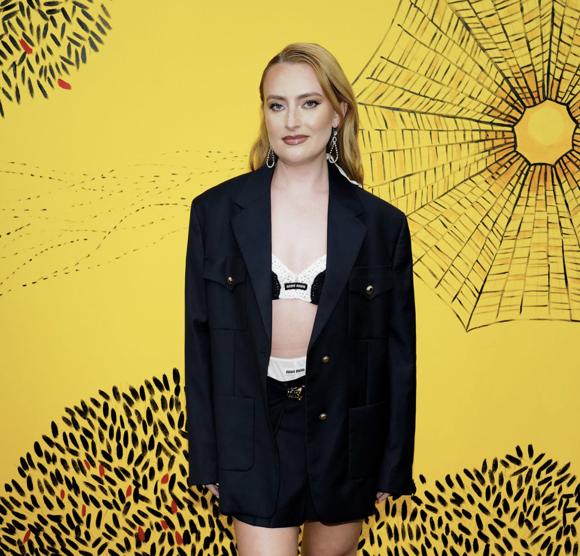 Woman in oversized blazer and bra top poses against a vibrant yellow backdrop with stylized leaf and sun illustrations.