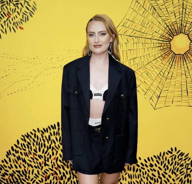 Woman in oversized blazer and bra top poses against a vibrant yellow backdrop with stylized leaf and sun illustrations.