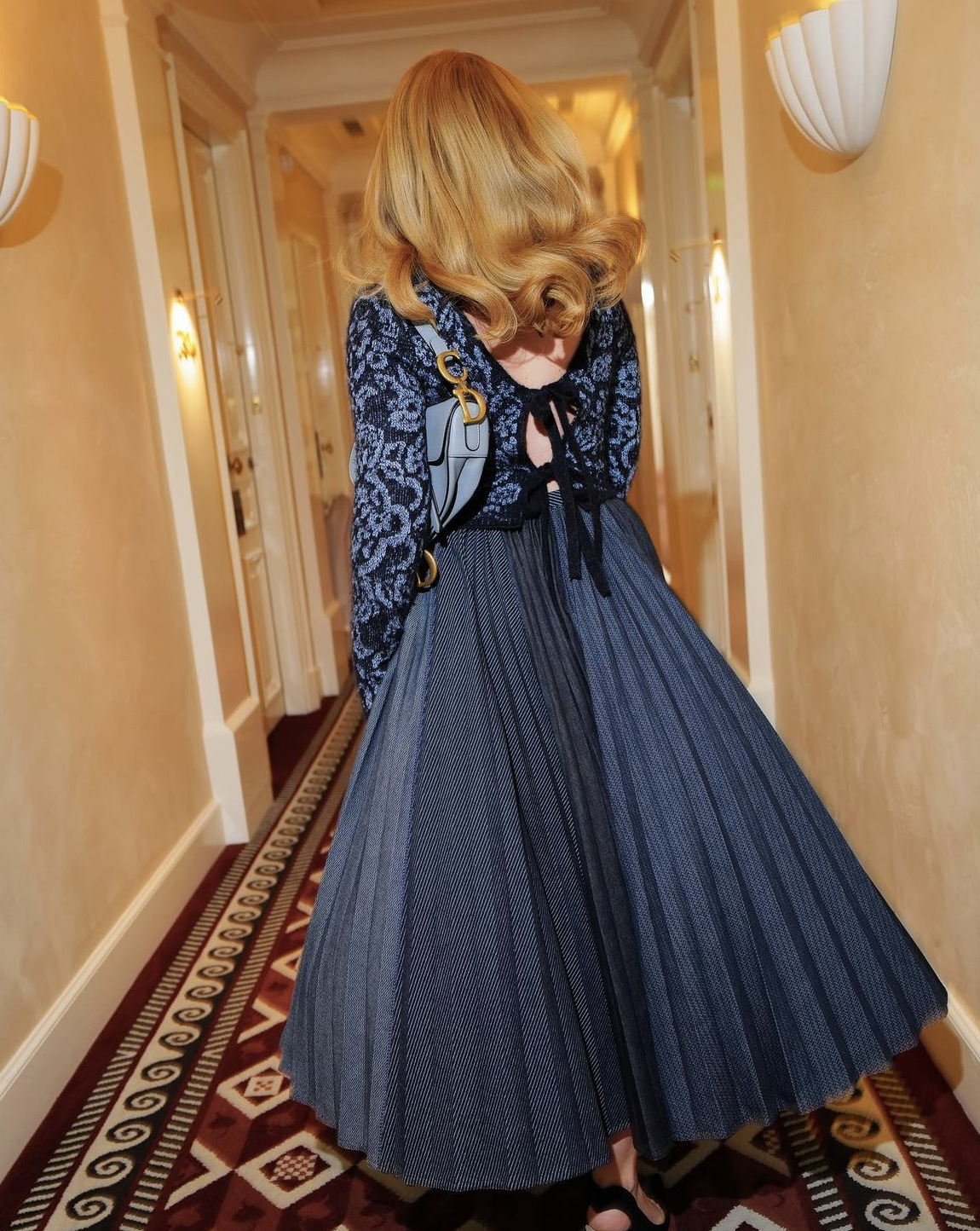 Woman in blue floral top and pleated skirt stands in hallway with patterned carpet and sconces, back to the camera, interior shot.