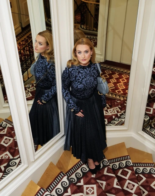 A poised woman in a blue patterned top and long pleated skirt stands on patterned stairs next to a mirror, indoors.