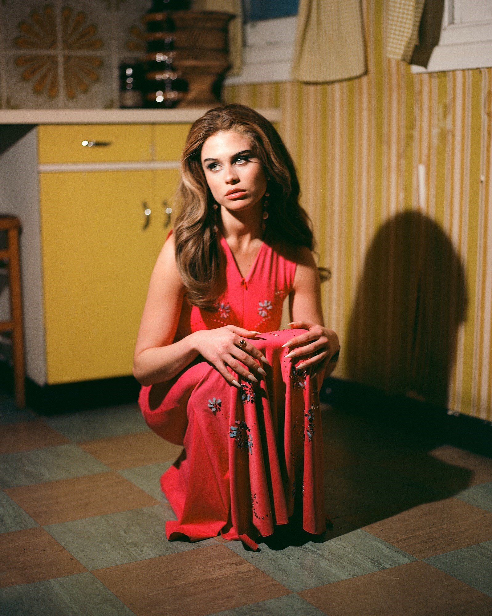 Young woman with long brown hair, wearing a pink dress, crouching on a checkered floor in an indoor setting with yellow cabinets.