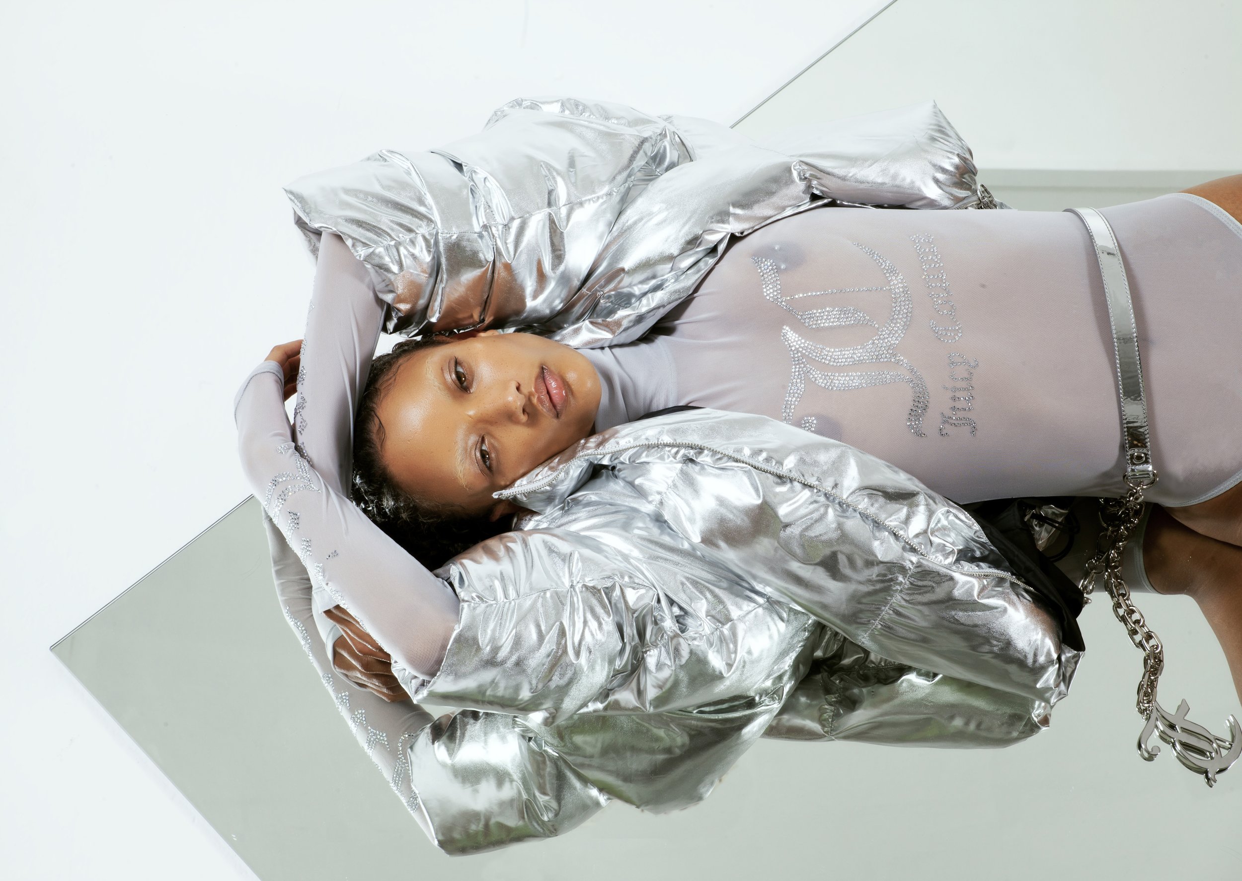 A woman lies on a mirrored surface wearing a silver jacket and bodysuit, with the 'Juicy Couture' logo prominently displayed.