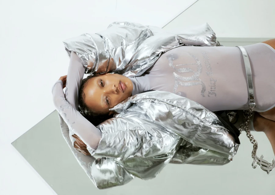 A woman lies on a mirrored surface wearing a silver jacket and bodysuit, with the 'Juicy Couture' logo prominently displayed.