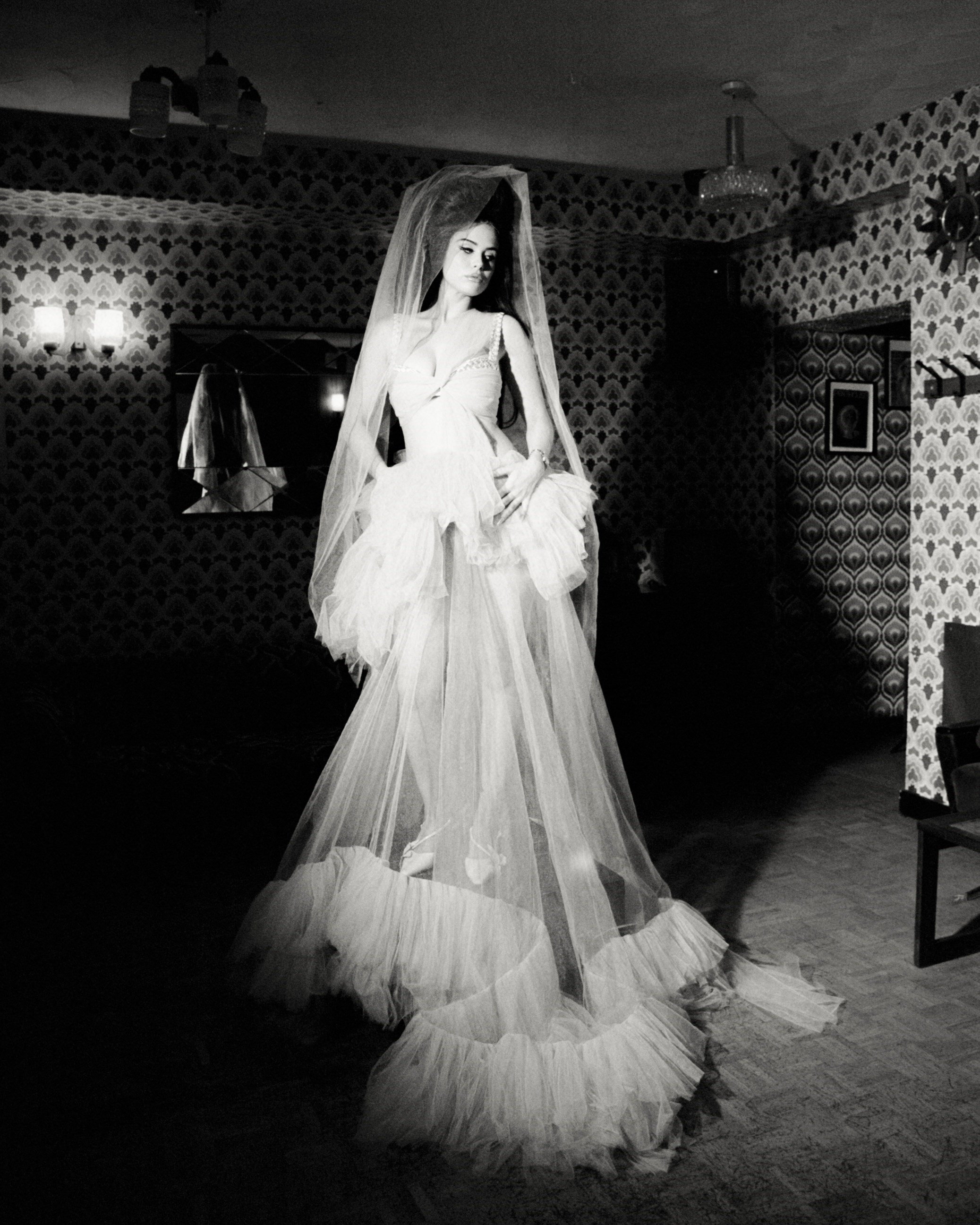 A woman in a white wedding dress and veil stands in a room with patterned wallpaper and dim lighting, looking at the camera.