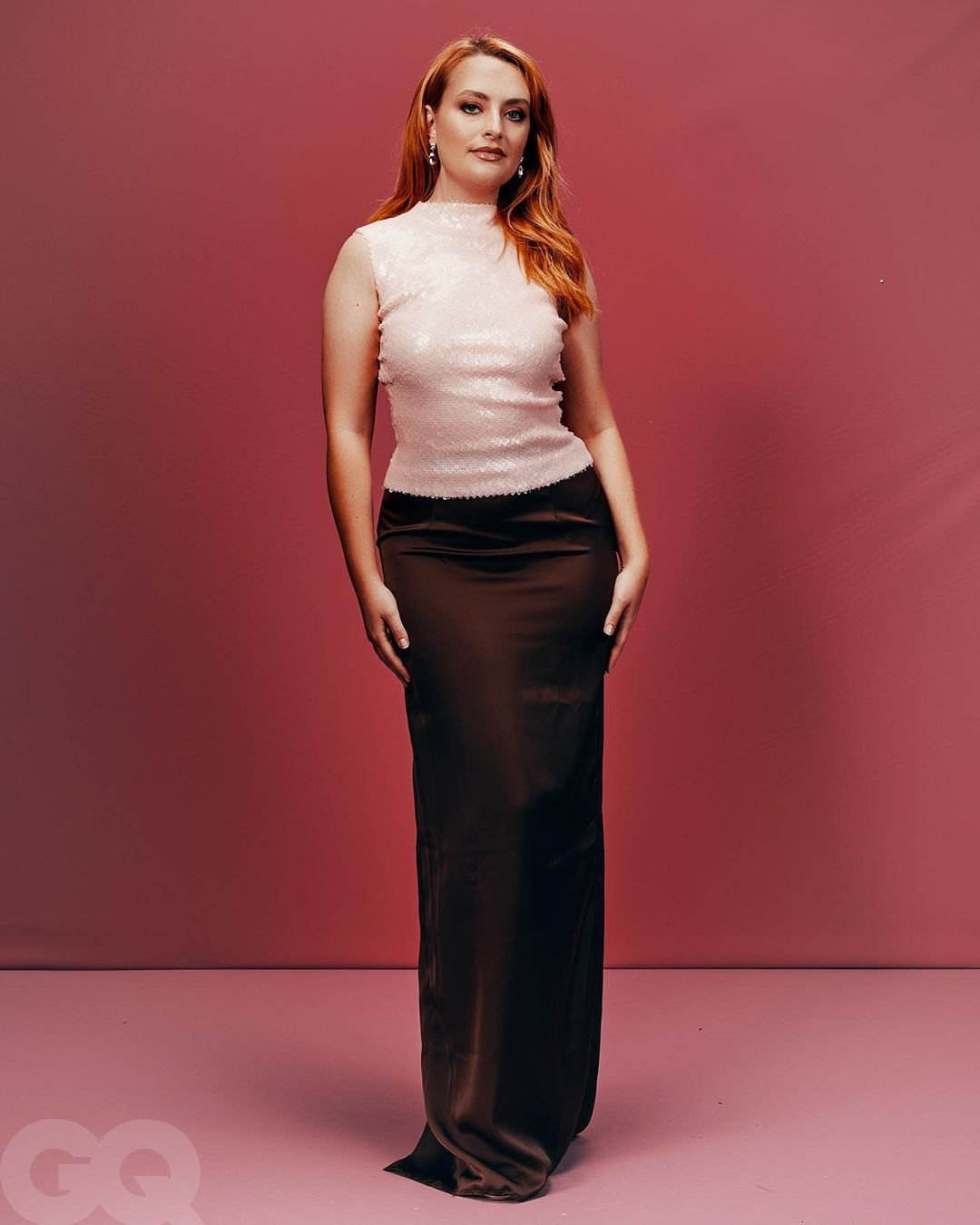 Woman in long brown satin skirt and sleeveless top, posing in front of a pink background, full-length portrait.