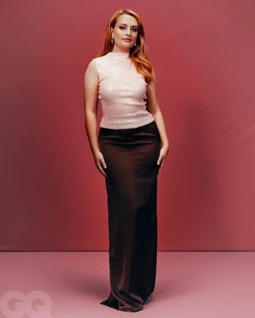 Woman in long brown satin skirt and sleeveless top, posing in front of a pink background, full-length portrait.