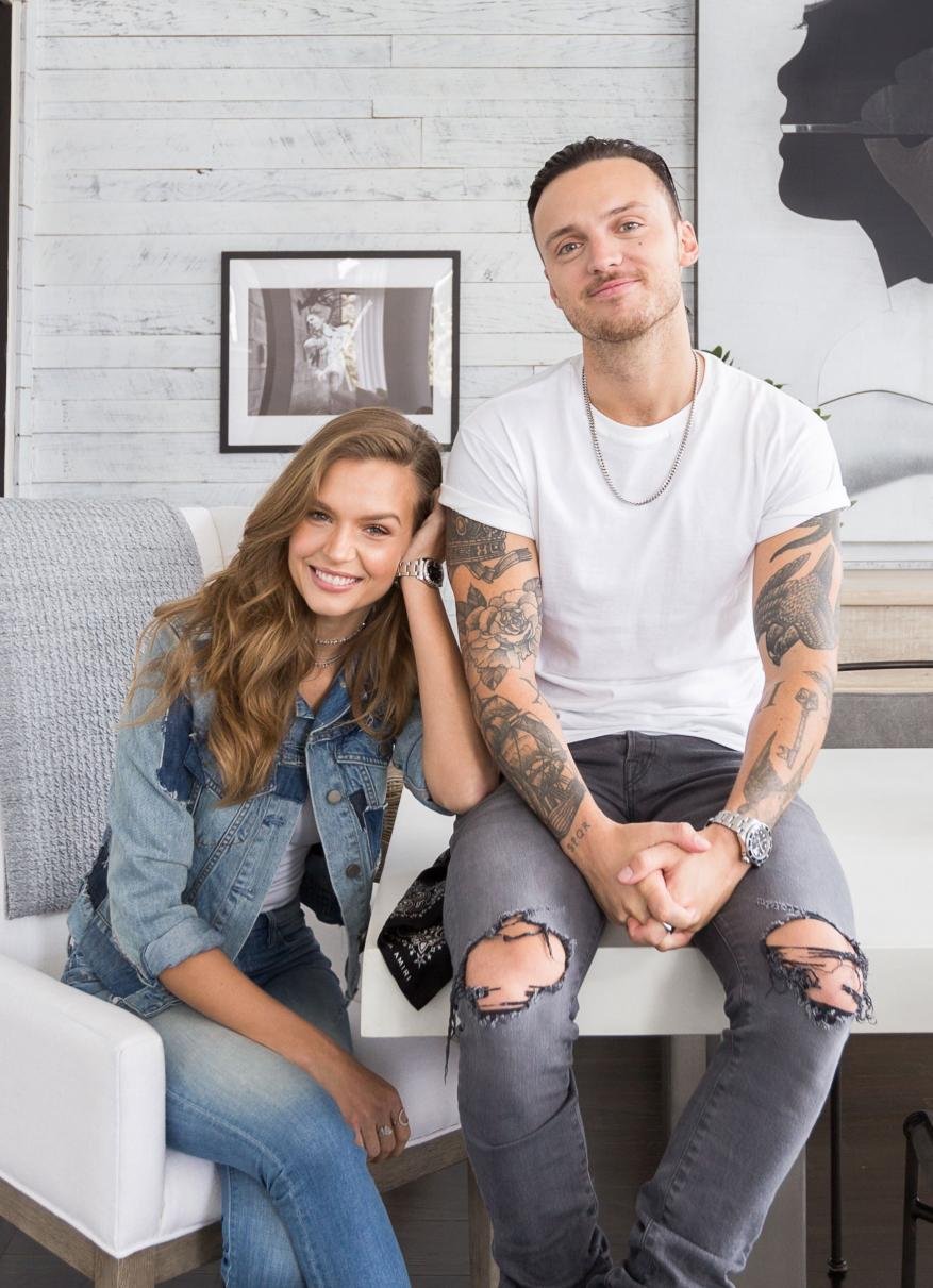 Indoor portrait of a smiling couple. The tattooed man is sitting on a white counter while the woman leans on the back of a white chair.