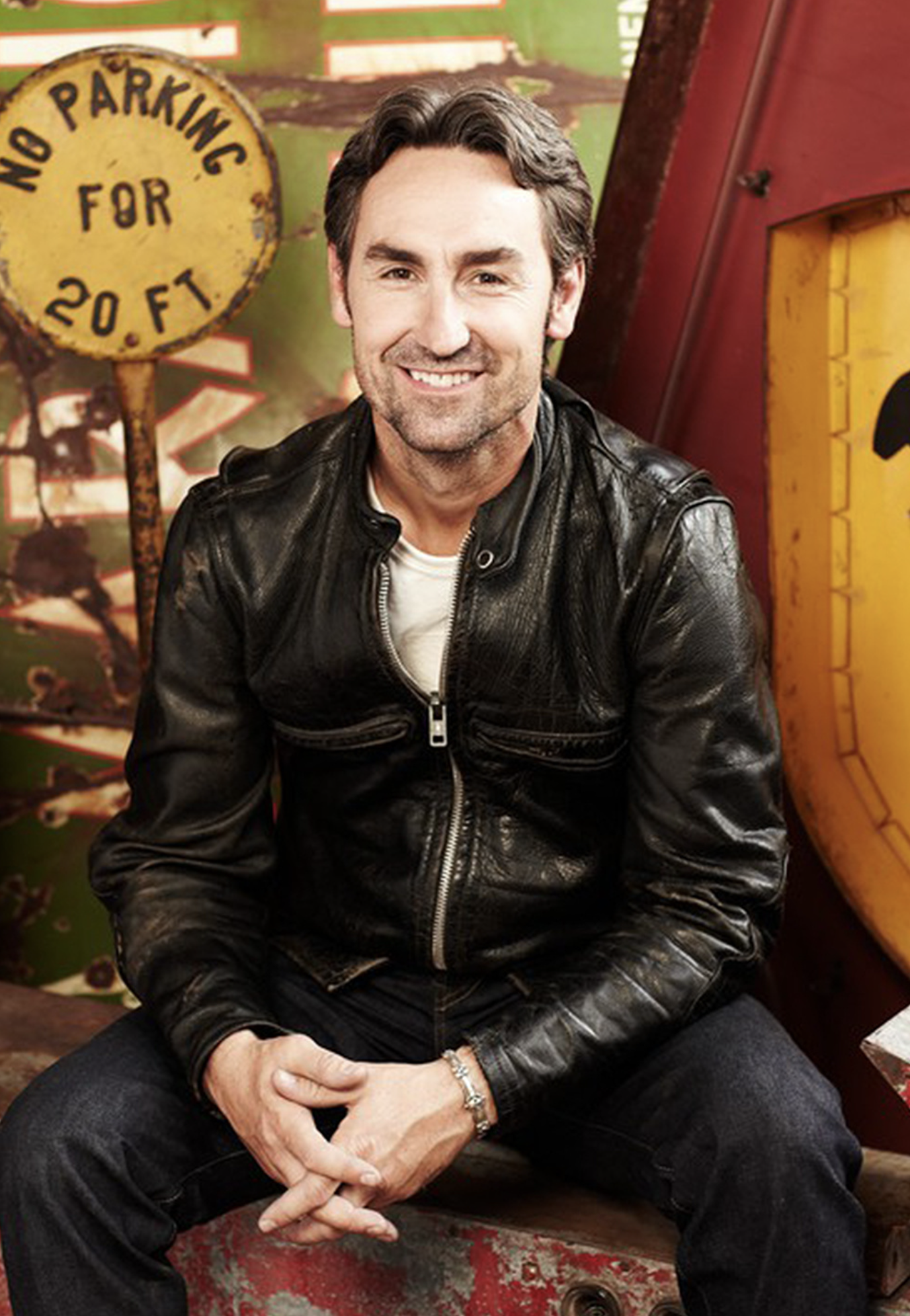 Portrait of a smiling man with dark hair, wearing a black leather jacket and jeans, sitting in front of a vintage industrial background.