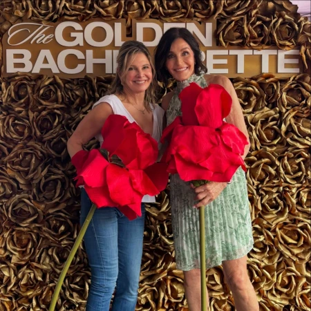 Two adult women pose with large red rose props in front of a floral background and "The Golden Bachelorette" text for event photography.