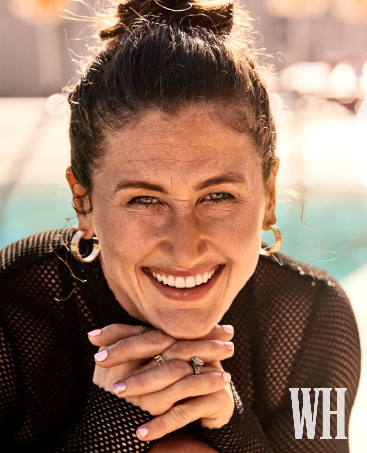 Close-up portrait of a smiling adult woman with hoop earrings and a black top, posing outdoors with natural lighting and a blurred background.