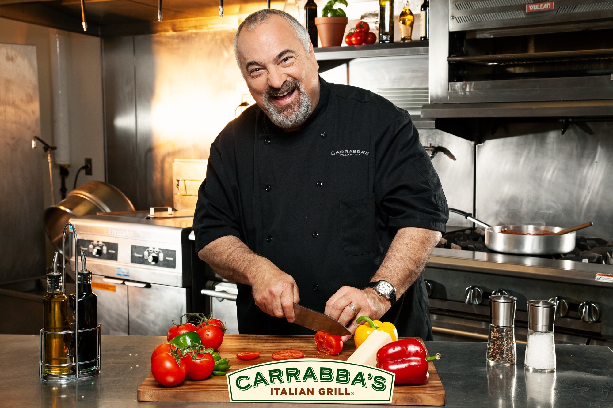 A chef in a restaurant kitchen smiles while slicing tomatoes on a wooden cutting board with colorful bell peppers nearby.