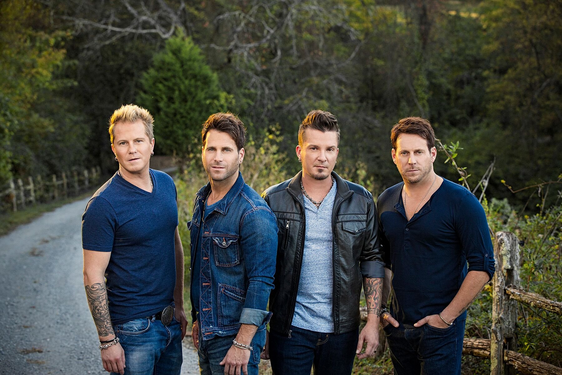 Group portrait of four adult men posing outdoors on a rural road, wearing casual clothing, with a forested background and soft lighting.