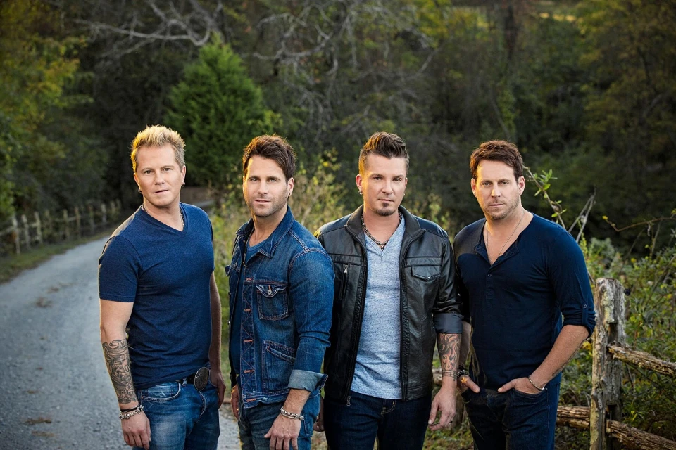 Group portrait of four adult men posing outdoors on a rural road, wearing casual clothing, with a forested background and soft lighting.