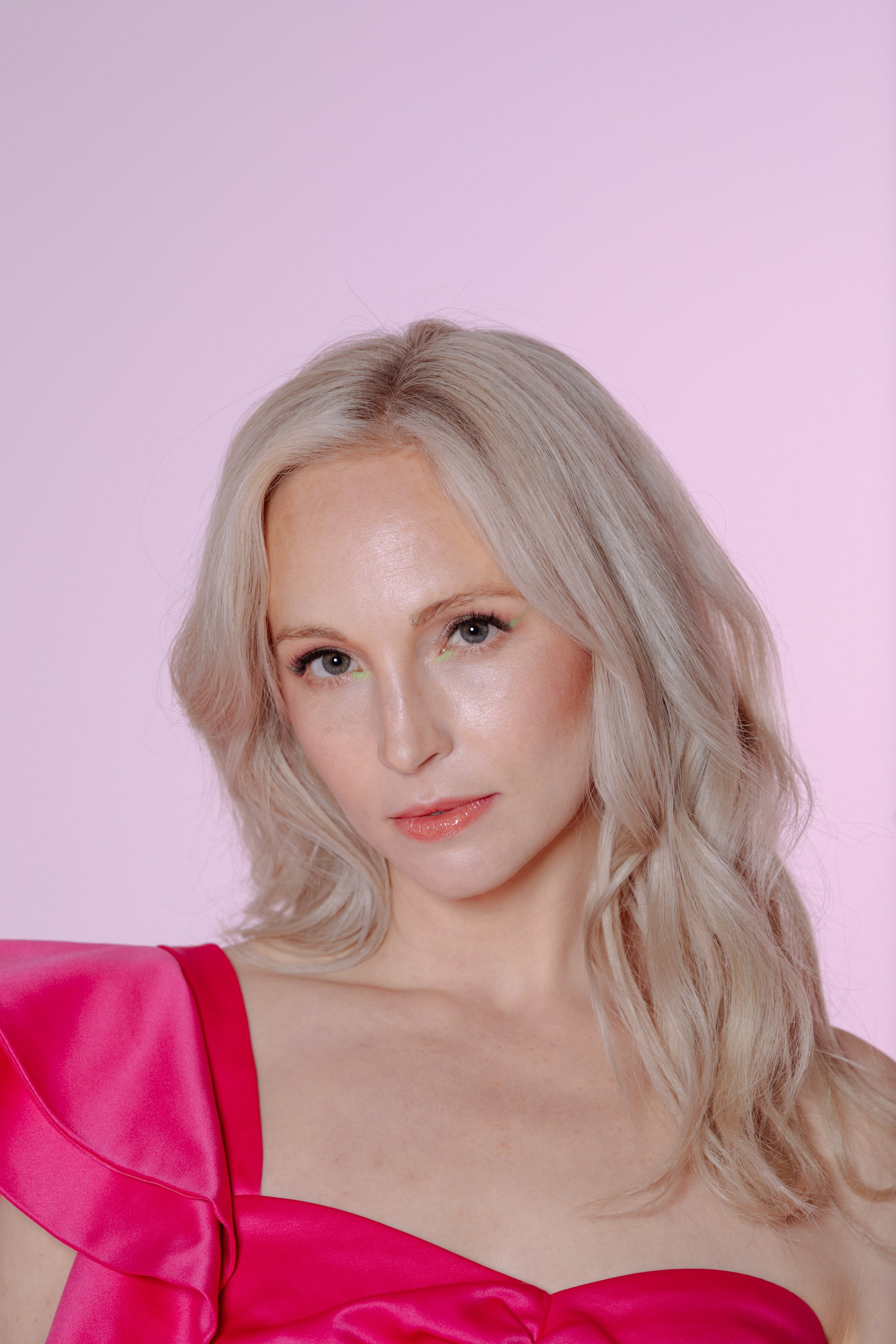 Blonde woman in a pink dress strikes a confident pose indoors under soft lighting, highlighting her elegant features and vibrant attire.