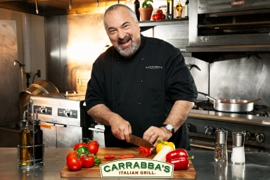 A smiling male chef in a black chef coat chopping tomatoes on a wooden cutting board in a professional kitchen.
