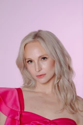 Portrait of a blonde woman wearing a vibrant pink satin top against a soft pink seamless background.