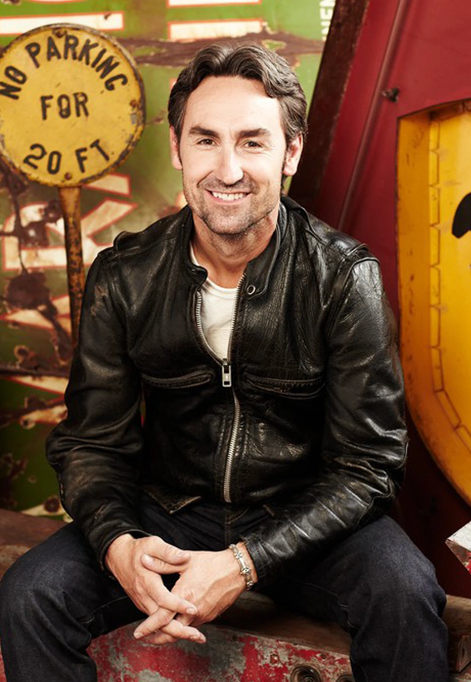 Man with dark hair wearing a black leather jacket and dark jeans seated against a backdrop featuring old parking signs.