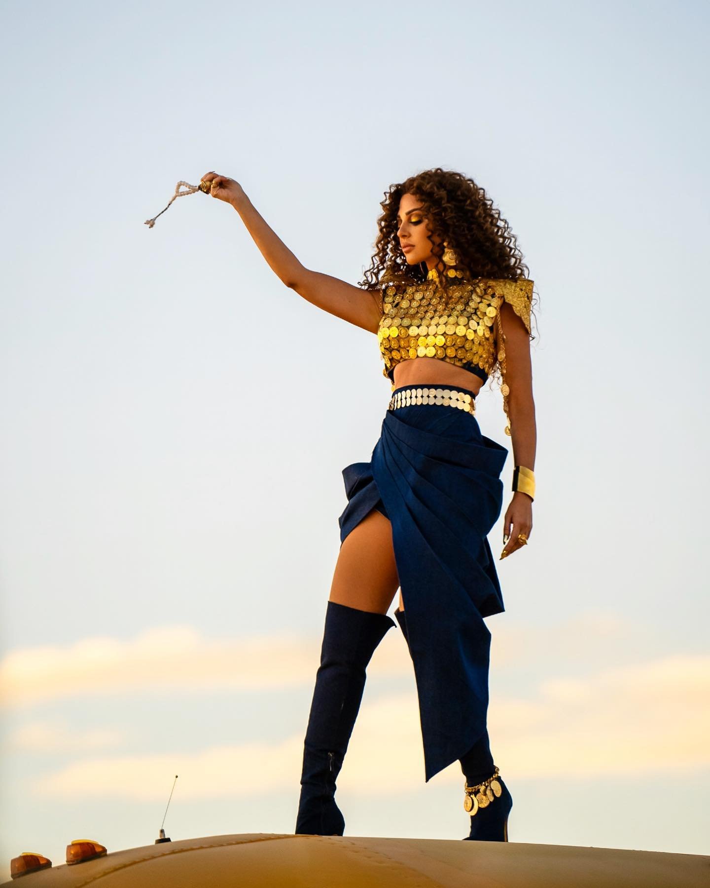 Adult woman wearing gold sequin crop top, navy skirt, and black boots stands on structure with a sky background in outdoor setting.