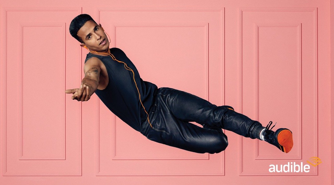Man in black tank top and leather pants floating mid-air with pink wall background, wearing orange earbuds and black and orange sneakers.