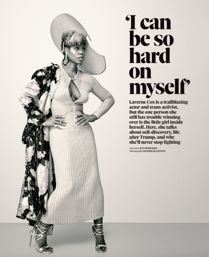 Laverne Cox poses indoors wearing a beige ribbed dress, silver heels, and a large hat, with a black and white patterned coat.