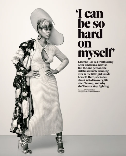 Laverne Cox poses indoors wearing a beige ribbed dress, silver heels, and a large hat, with a black and white patterned coat.