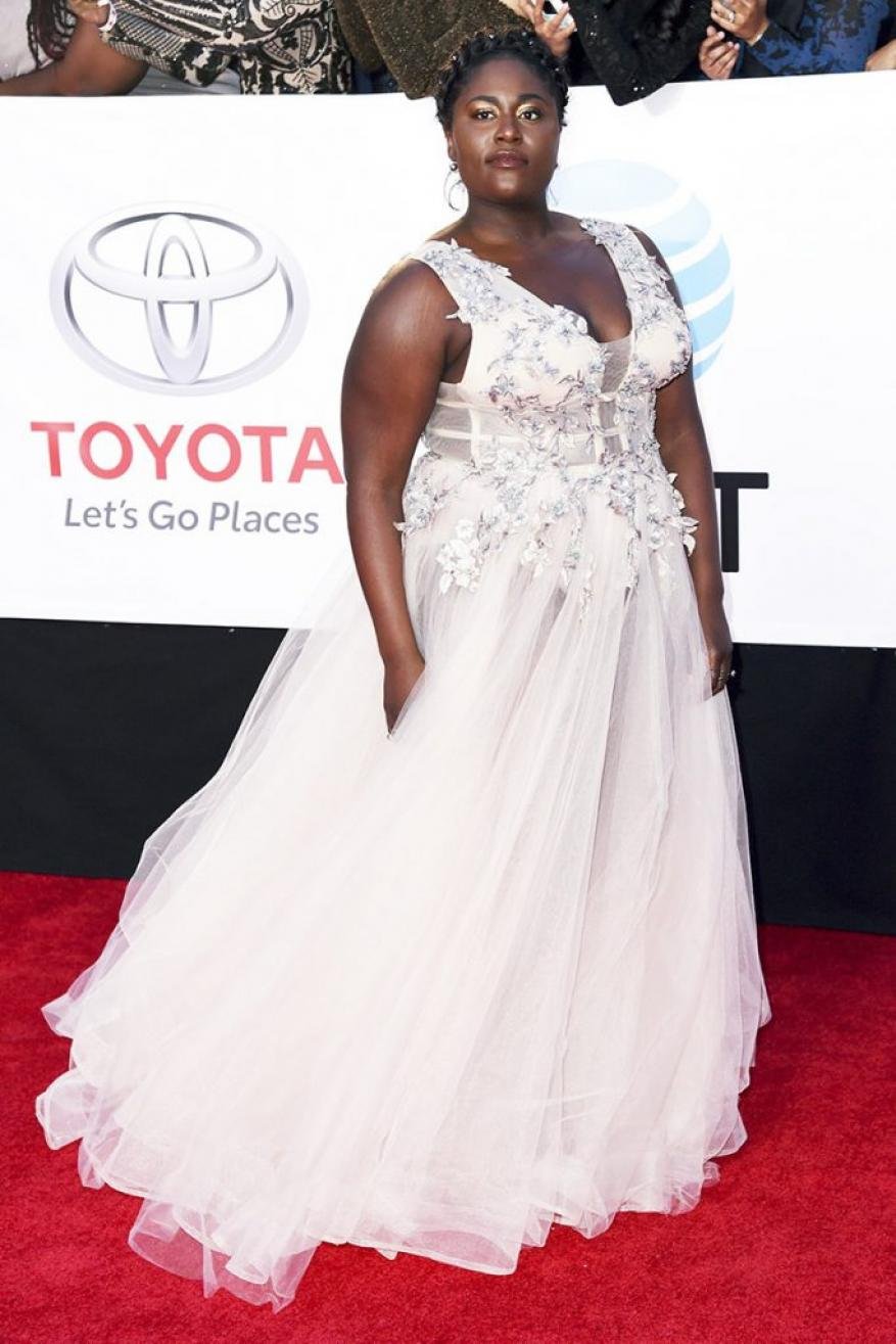Woman on red carpet in a formal gown with beaded detail, standing in front of a branded backdrop at a red-carpet event.