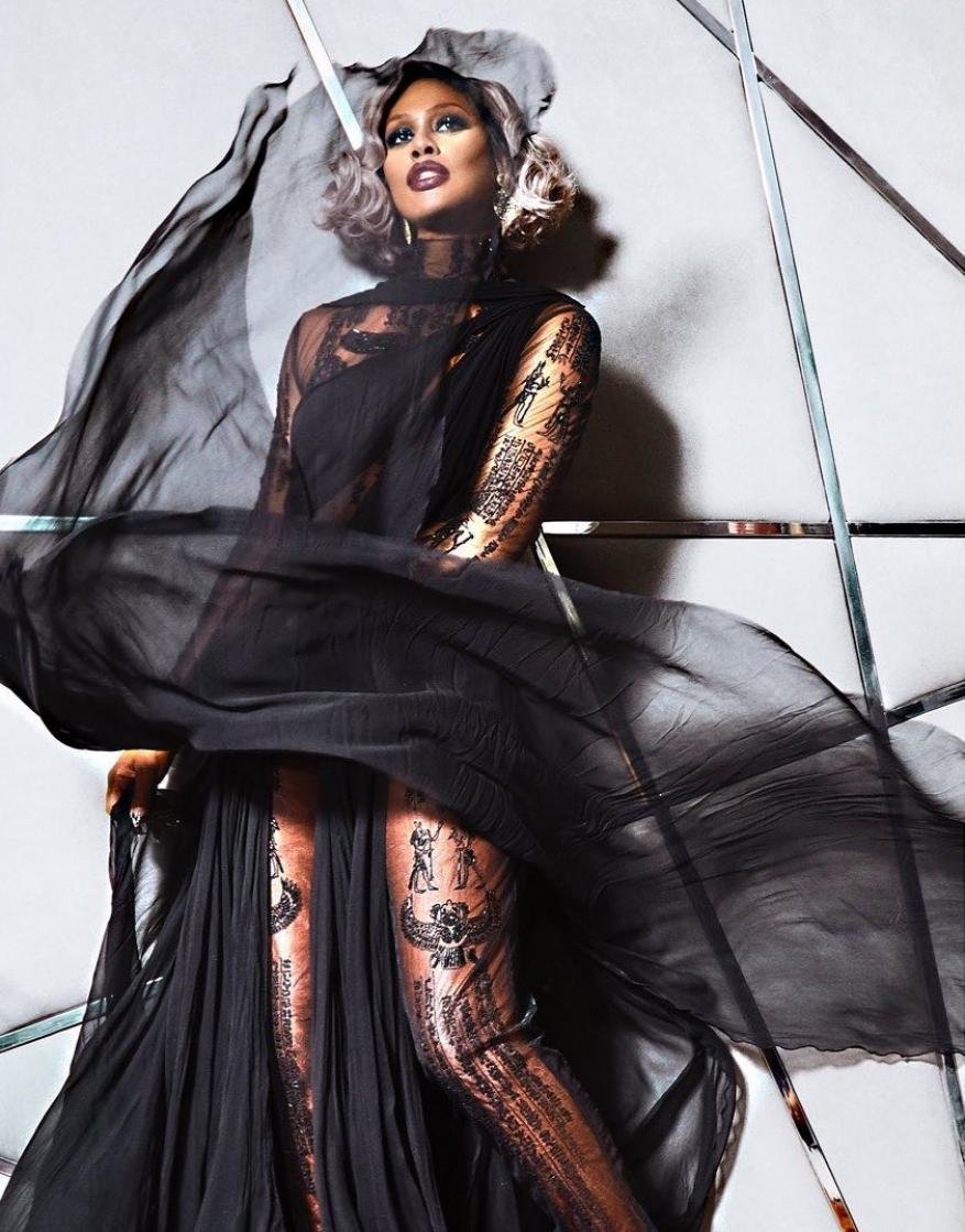 Fashionable woman poses indoors wearing a black gown and sheer shawl, with soft blonde hair and striking makeup for a fashion shot.