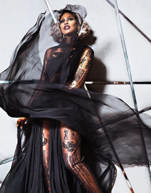 Fashionable woman poses indoors wearing a black gown and sheer shawl, with soft blonde hair and striking makeup for a fashion shot.