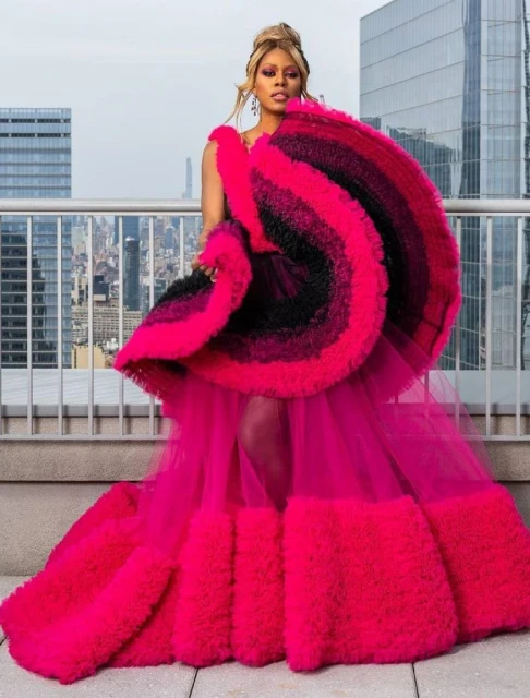 Elegant woman poses in a hot pink gown with a ruffled overlay on a rooftop overlooking a city skyline. High-fashion editorial photo.