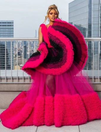 Elegant woman poses in a hot pink gown with a ruffled overlay on a rooftop overlooking a city skyline. High-fashion editorial photo.