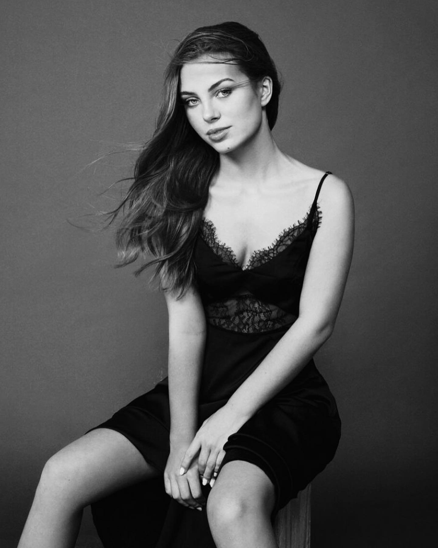 Monochrome portrait of a young woman with long brown hair sitting, wearing a black lace-trimmed slip dress, with a solid gray background.