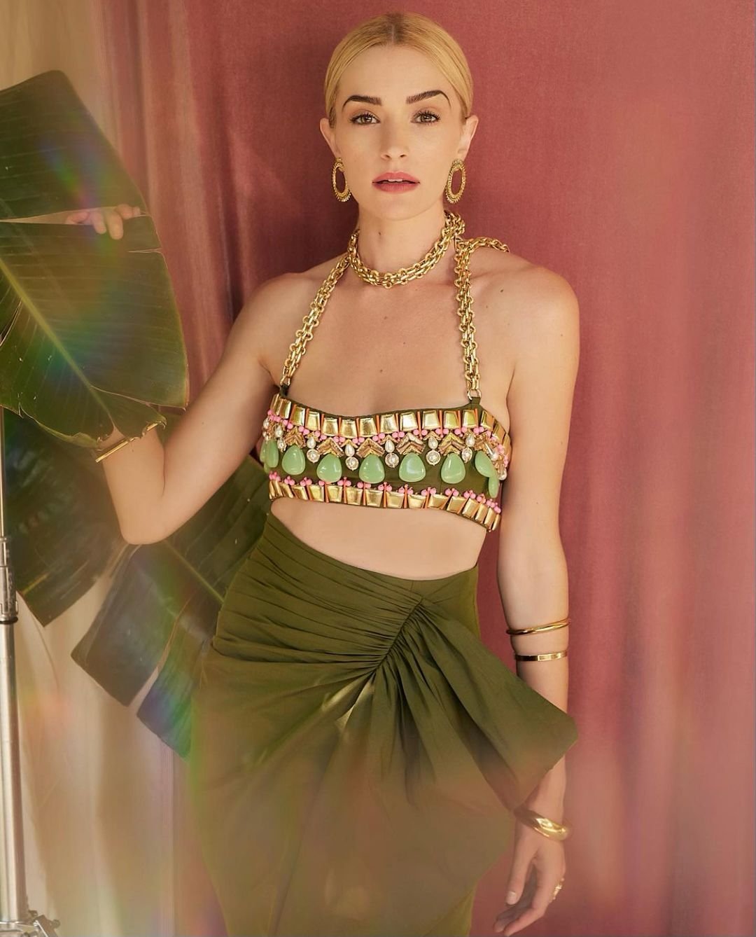 A fair-skinned woman with blonde hair poses in an olive green skirt and beaded halter top in front of a pink backdrop with a banana leaf.