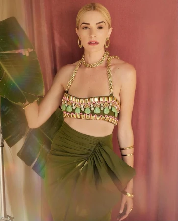A fair-skinned woman with blonde hair poses in an olive green skirt and beaded halter top in front of a pink backdrop with a banana leaf.