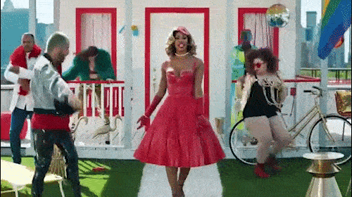 Adult female in red dress poses with party guests on outdoor deck with disco ball, bike, and colorful cityscape background.