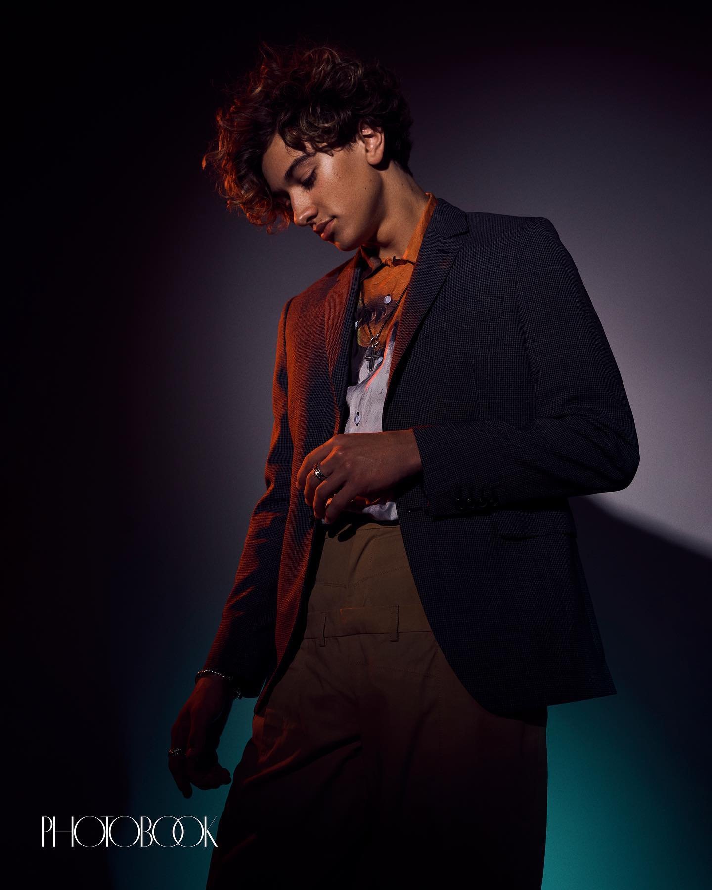 Portrait of a young adult male in a dark blazer and light brown pants looking down in an indoor setting with colored lighting.