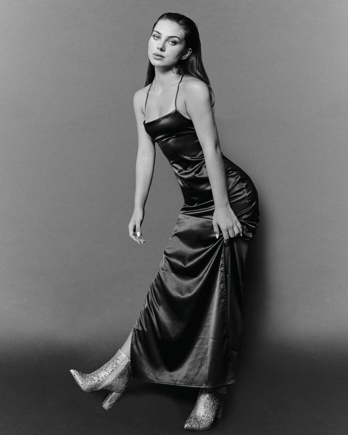 Monochrome studio portrait of a young woman in a satin dress and glitter boots, posing and looking at the camera with a neutral expression.